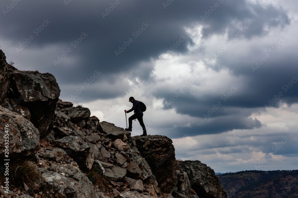Obraz premium Man hikes up rocky mountain with walking sticks under cloudy sky. Person climbs steep terrain, carries backpack, explores nature wilderness. Dramatic storm clouds gather above summit.