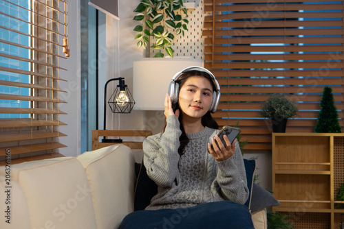 A happy Asian woman wears wireless headphones and holds her smartphone while relaxing on the sofa.