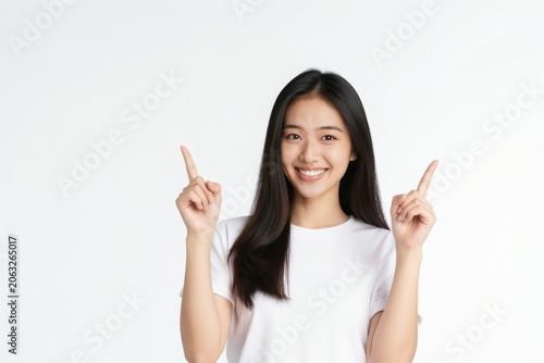 Smiling young Asian woman in white shirt points fingers up. She gestures to present information or indicate a direction against a plain background. Copy space available for text.