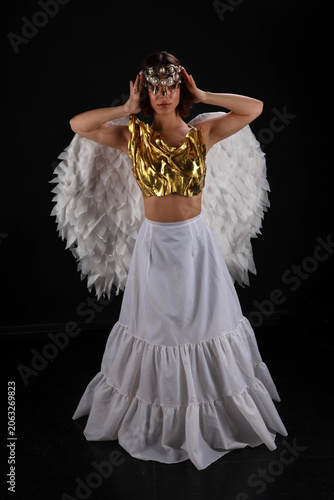 Full length cinematic portrait of a beautiful young woman in a golden corset and long white skirt with angel wings on a black background
