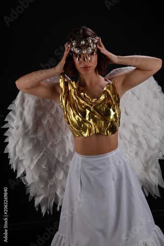 Medium shot of a beautiful young woman in a golden corset and white skirt with angel wings against a black background