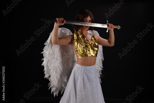 Medium shot of a beautiful young woman in a golden corset and white angel wings holding a medieval sword against a black background