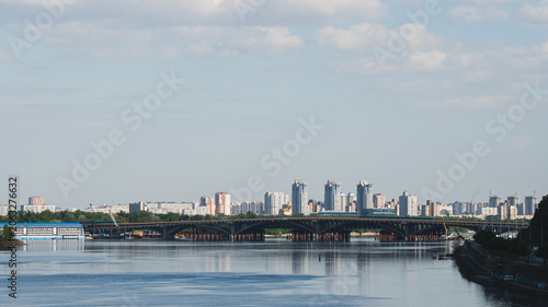 Kyiv city skyline view across the Dnieper River featuring the Metro Bridge, water reflections, and modern residential buildings under a cloudy sky in Ukraine.
