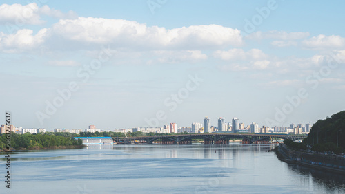 Kyiv city skyline view across the Dnieper River featuring the Metro Bridge, water reflections, and modern residential buildings under a cloudy sky in Ukraine.