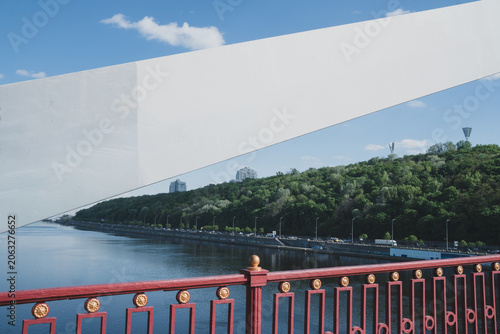 Scenic city view of Kyiv, Ukraine, from the Park Bridge over the Dnieper River, showing Naberezhne Highway, green hills, and red bridge railings under a blue sky.