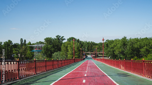 Pedestrian and bicycle bridge with red railings and a dedicated red cycling path leading towards a green park under a clear blue sky on a sunny day.