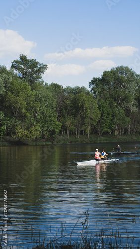 Distant view of rowers training on a calm river with green forest background, featuring a double scull and a single scull boat.