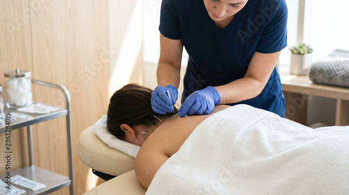 A young Asian woman receives acupuncture treatment from a Caucasian female practitioner in a serene clinic environment. The practitioner wears gloves and focuses on the woman's back.