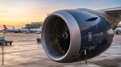 Close-up of a jet engine on an aircraft at sunset. The engine is detailed with visible components and textures. Airport background with parked planes.
