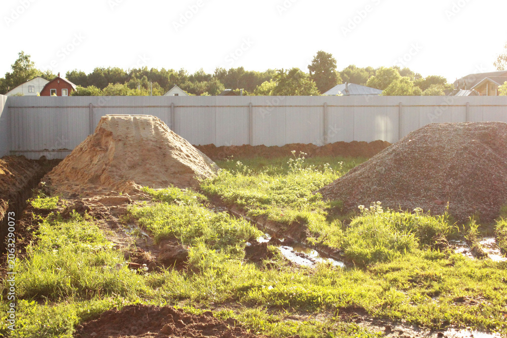 Obraz premium A sunlit construction site shows large soil piles, muddy ground, and fresh excavation work inside a fenced residential area, with trees and houses visible in the distance.
