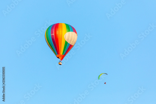 Masovian Voivodeship, Poland - 07 August 2018: View of a colorful rainbow-striped hot air balloon and a paraglider flying in a clear blue sky.