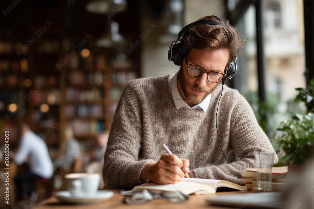 Fototapeta premium A young man journaling with headphones on in a coffee shop. 