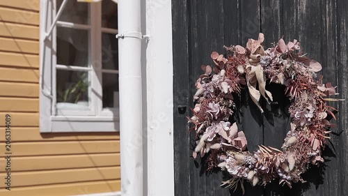 charming, close-up detail of a wooden building in the old town of Porvoo. A rustic floral wreath hangs on a weathered black door, while a bright yellow wooden house with a striped awning is softly