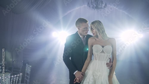 Bride and groom embrace during first dance at wedding reception, illuminated by dramatic blue spotlights and chandelier, romantic atmosphere, elegant white gown and dark suit, soft focus background