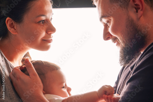 Happy young family playing and kissing newborn baby and smiling and hugging at home, loving family holding their son in arms.