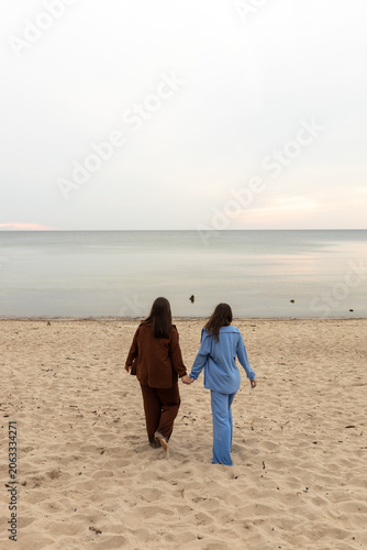 Two girlfriends walk hand in hand along a sandy beach towards the sea.
