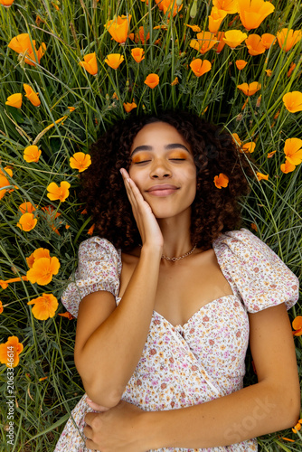 A young woman with curly hair lies peacefully in orange wildflowers, wearing a floral dress, embodying relaxation and sunny happiness. Generative AI.