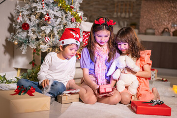 Children in Santa hats and reindeer antlers gather by a glowing decorated Christmas tree opening gifts at home.