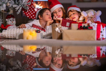 Excited children in Santa hats open Christmas presents together by a decorated tree and cozy fireplace at home.