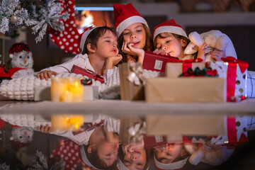 Excited children in Santa hats open Christmas presents together by a decorated tree and cozy fireplace at home.