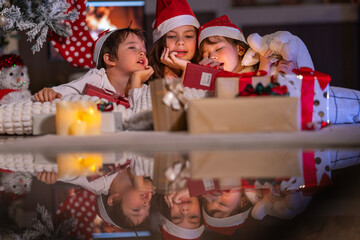 Excited children in Santa hats open Christmas presents together by a decorated tree and cozy fireplace at home.