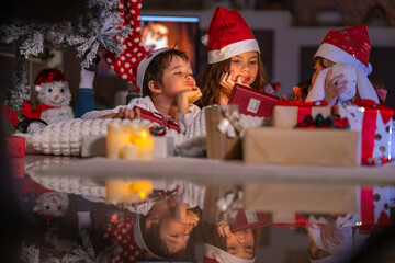 Excited children in Santa hats open Christmas presents together by a decorated tree and cozy fireplace at home.