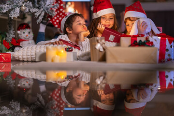 Excited children in Santa hats open Christmas presents together by a decorated tree and cozy fireplace at home.