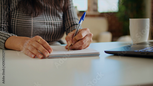 Tableau sur toile Teleworker at desk transcribing data from laptop to notepad, finalizing project, remote worker solving job tasks before deadline, writing info on paper using pen, close-up panning shot