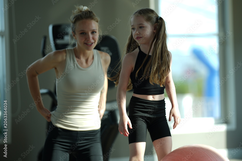 Naklejka premium Mother and young daughter stretch and exercise together on a mat in a bright gym, sharing a healthy active lifestyle.