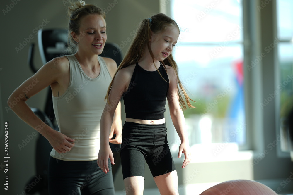 Naklejka premium Mother and young daughter stretch and exercise together on a mat in a bright gym, sharing a healthy active lifestyle.
