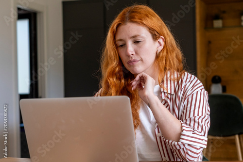 Konstfotografi Red-haired woman working on a laptop at a home office desk, focused on her scree