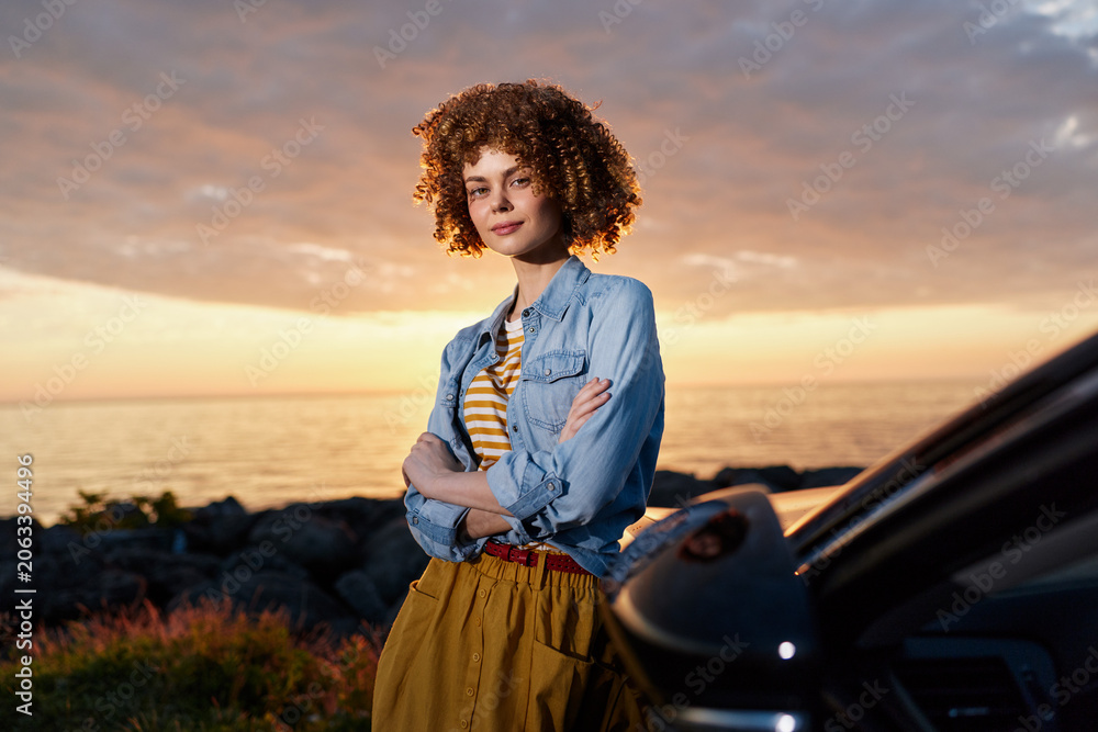 Fototapeta premium Confident woman with curly hair leaning against a car at sunset, wearing a denim shirt and striped top. Lifestyle portrait showcasing outdoor style, independence, and warm, relaxed mood.