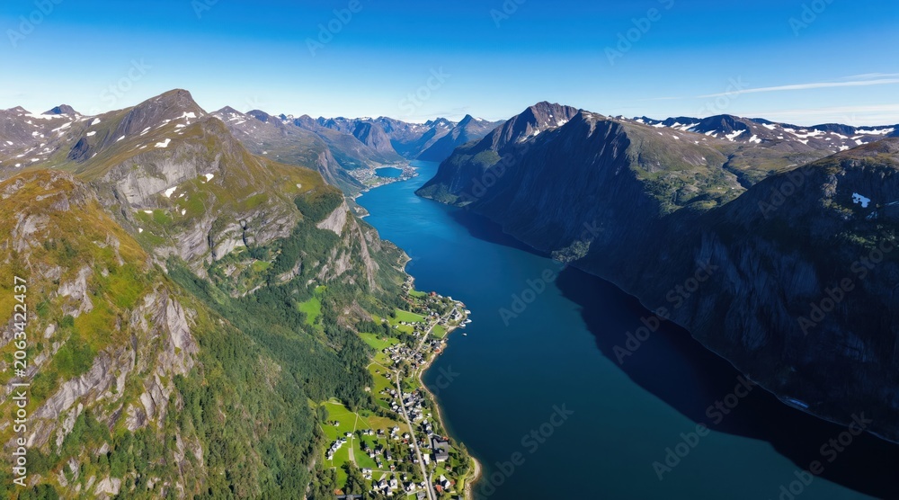 Fototapeta premium Aerial view of deep blue fjord between tall green mountains. Small village rests on green shore beside calm water. Bright clear blue sky covers peaceful natural landscape.