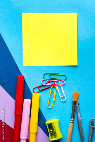 Blank yellow sticky note on colorful blue background with markers paperclips paintbrush compass and sharpener back to school copy space. 