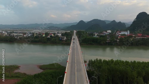 Bridge Crossing Calm River Into Small Town, Vehicles Moving Toward Clustered Houses With Karst Hills Beyond, Soft Dusk Glow Over Water And Settlement In Phong Nha Vietnam