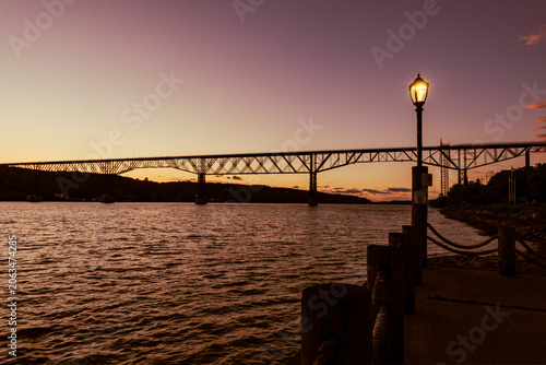 The Walkway Over the Hudson captures a stunning sunset. The historic bridge spans across the wide river. Soft purple and orange hues paint the evening sky. Glowing vintage lamppost lights the walkway.