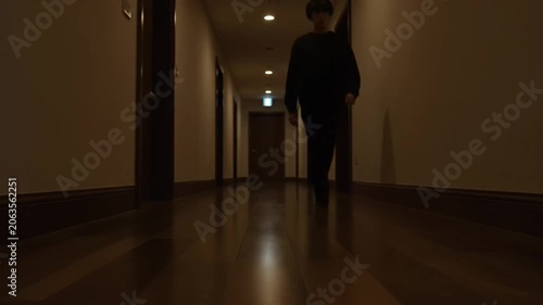 A man walks down a dark hotel corridor at night.