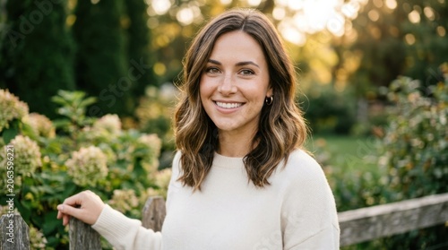 A woman with shoulder-length brown hair, wearing a white sweater, standing in a garden with greenery and flowers.
