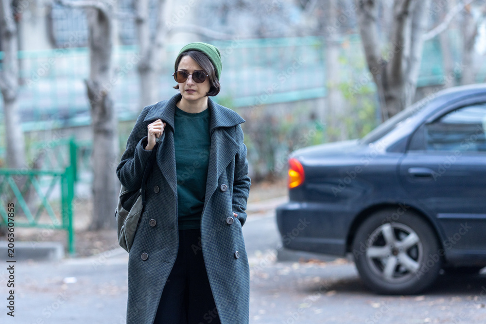 Fototapeta premium Portrait of a woman wearing a dark coat, green beanie, and sunglasses standing outdoors with a car in the blurred background.