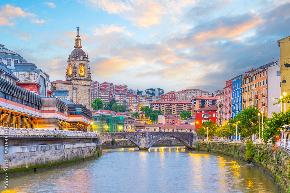 Obraz premium The historic Ribera Market and colorful waterfront buildings reflected in the Nervion River, Bilbao, Spain.