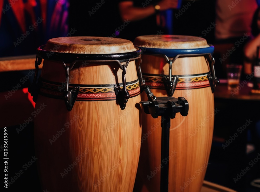 Fototapeta premium Pair of conga drums with detailed wood grain sit ready for music performance. Traditional percussion instruments await rhythm in dimly lit bar setting. Colorful band adorns drums for vibrant salsa