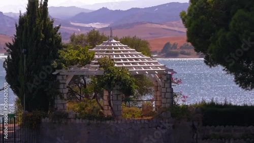 Drone footage drifting sideways with telephoto compression beside El Chorro's ornate colonnade, skimming flowering urns, tall cypress, and a shimmering inlet backed by burnt-ochre plains.