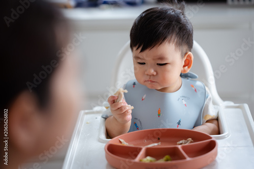 Asian Baby Using Finger to Feed Herself a Food with Baby Led Weaning Concept.Cute Asian baby boy eating finger food in high chair.	
