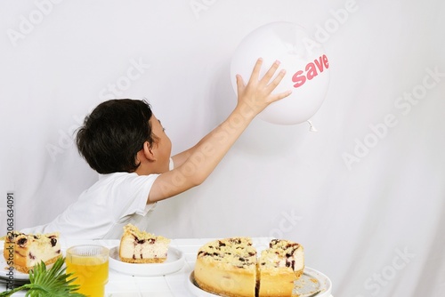 Cute little boy reaching for white balloon beside cakes and juice in bright studio with playful lifestyle and celebration concept.