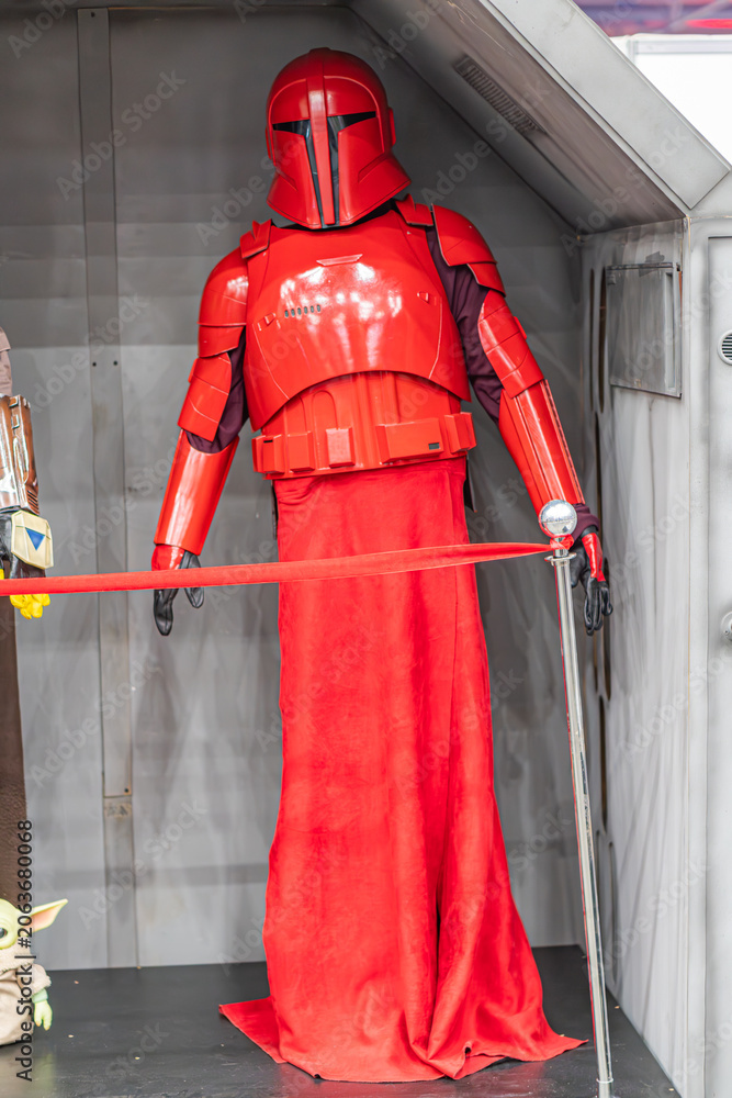 Naklejka premium Life-sized Star Wars costume figure in red armor, against a dark backdrop Red cape, helmet with visor, chest piece adorned with symbols, articulated arm sections, b - Kyiv, Ukraine - June 7, 2025