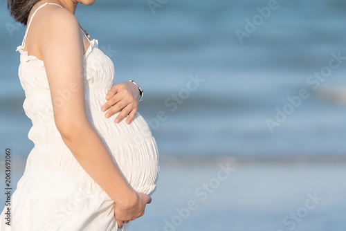 Pregnant woman in white dress gently placing her hands on her belly by the sea, reflecting the beauty and health of pregnancy.