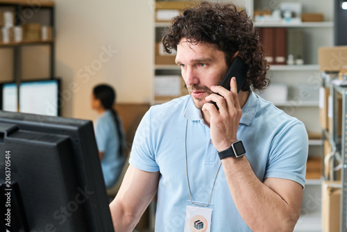 Quadro su tela Medium shot of young man managing order pickup point, speaking on smartphone and checking monitor