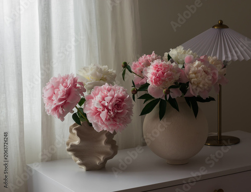 Bouquets of pink peonies in ceramic vases and a table lamp on a white table