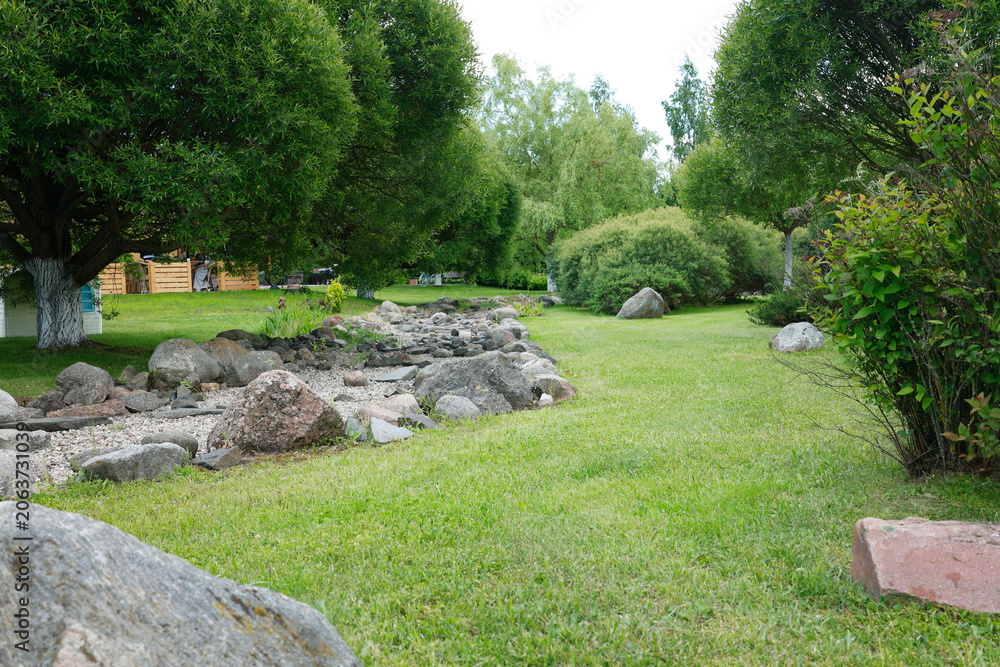 Fototapeta premium Beautiful rock garden with decorative natural stones on green grassy lawn. Professional landscaping design featuring dry creek bed with large boulders surrounded by lush green trees. Scenic