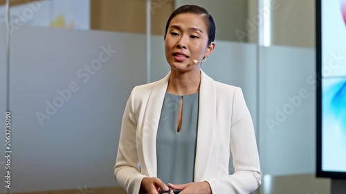 Confident Asian businesswoman in white blazer delivers a corporate briefing with headset and presentation remote, honoring business women's day inside a bright glass-walled executive meeting space